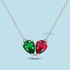 May include: A silver necklace featuring a pendant with two teardrop-shaped gemstones, one emerald green and one ruby red. The gemstones are set in a V-shaped silver setting, suspended from a delicate chain. The text "AAVRA" is visible.