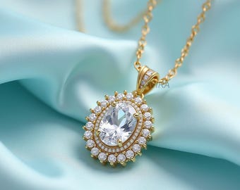 May include: A gold-toned necklace with a large, oval-shaped pendant. The pendant features a central, clear gemstone surrounded by smaller, clear stones. The necklace chain is also gold-toned, and the background is a light blue fabric.
