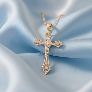May include: A gold-colored cross pendant necklace with a delicate chain. The cross features a central, large, clear gemstone and smaller gemstones on each arm. The pendant is displayed on a light blue, silky fabric.