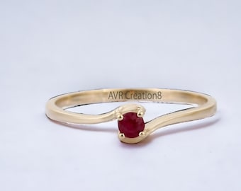 May include: A gold ring with a single red ruby gemstone set in the center. The band is thin and has a curved design.
