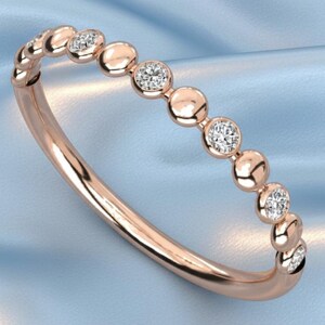 May include: Rose gold ring featuring a series of small, round, bezel-set diamonds. The band is a polished, thin metal. The ring is displayed on a light blue, silky fabric.
