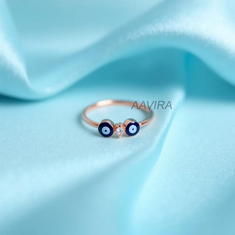 May include: Rose gold ring featuring two blue evil eye charms and a small clear stone in the center. The ring is set against a light blue satin background. The brand name "AAVIRA" is visible.