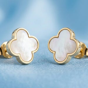 May include: A pair of gold-colored stud earrings. Each earring features a four-leaf clover design with a white, pearlescent center. The earrings are set against a soft, light blue background.
