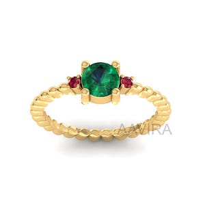 May include: A gold ring featuring a large, round emerald center stone flanked by two smaller ruby stones. The ring band has a textured, twisted rope design. The ring is set against a white background.