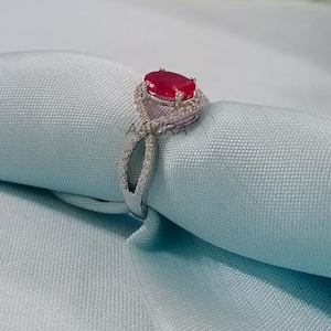 May include: A silver ring with a large oval ruby gemstone surrounded by smaller diamonds. The ring has a delicate, twisted band.
