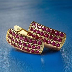 May include: A pair of gold hoop earrings with a row of red cubic zirconia stones. The earrings are on a blue background.