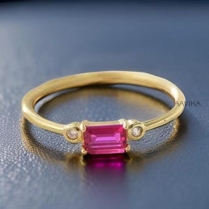 May include: A gold ring featuring a rectangular, vibrant pink gemstone at the center, flanked by two smaller, round, clear stones. The ring has a delicate band and a polished finish, set against a dark background.