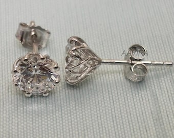 May include: A pair of silver stud earrings with a large, clear, round-cut gemstone. The earrings have a detailed, floral-inspired setting and a post-and-butterfly closure. The earrings are displayed on a light blue surface.
