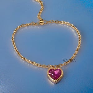 May include: A gold chain bracelet with a pink heart charm. The heart charm has a clear pink gemstone in the center.