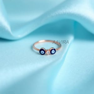 May include: Rose gold ring featuring two blue evil eye charms and a small clear gemstone. The ring is set against a light blue satin background. The brand name "AAVIRA" is visible on the right side of the ring.