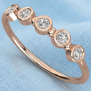 May include: A rose gold ring with five round, clear gemstones set in individual bezels. The ring is on a light blue fabric background. The gemstones are evenly spaced along the band.