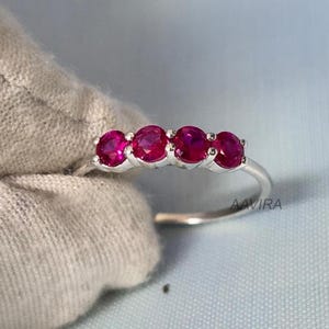 May include: A silver ring featuring a row of four vibrant, round, ruby gemstones. The rubies are a deep, rich red color, set closely together. The ring is displayed against a light blue background and a gray fabric.