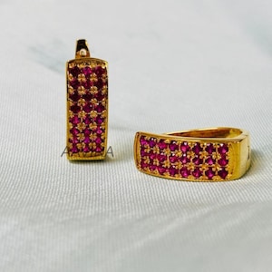 May include: A pair of gold-toned earrings with a rectangular design. Each earring is encrusted with numerous small, round, ruby-colored stones. The earrings are displayed on a light-colored surface.