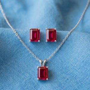 May include: A silver necklace and matching stud earrings set. The jewelry features rectangular, deep red gemstones set in silver. The necklace pendant and earrings are displayed on a blue fabric background.