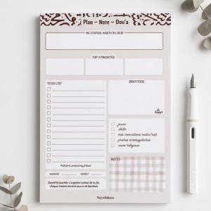 Peut inclure: Un bloc-notes gris clair avec des sections pour la planification quotidienne, comprenant une liste de tâches, la gratitude et des notes. Le bloc-notes a une bordure décorative et du texte en arabe. Un stylo plume blanc est à côté.