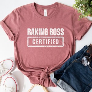 May include: A dusty rose t-shirt with the text "Baking Boss Certified" in white lettering. The shirt is tied in a knot at the front and is displayed with a pair of white sneakers, a wooden bead necklace, and a pair of blue denim shorts.