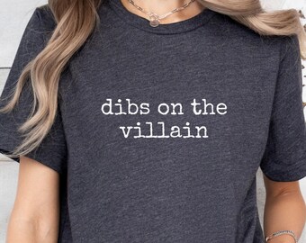 Könnte beinhalten: Dunkelgraues T-Shirt mit dem weißen Aufdruck "dibs on the villain". Das T-Shirt hat kurze Ärmel und einen Rundhalsausschnitt. Der Text ist mittig auf der Brust platziert.