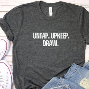 May include: Dark gray t-shirt with the text "UNTAP. UPKEEP. DRAW." in white. The shirt is paired with light blue denim jeans and white canvas sneakers with red accents. The items are arranged on a light wood surface.