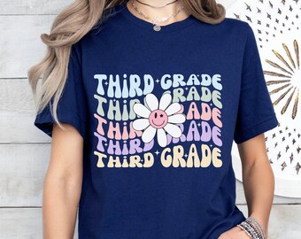 Op de afbeelding: Een marineblauw T-shirt met een kleurrijke afbeelding van een madeliefje met een smiley-gezicht en de tekst "THIRD GRADE" die in een regenboogpatroon wordt herhaald.