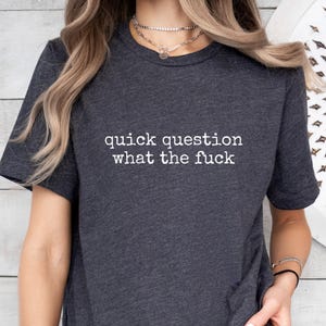May include: Dark gray t-shirt with the phrase "quick question what the fuck" printed in white. The shirt has short sleeves and a crew neck.
