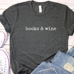 Könnte beinhalten: Dunkelgraues T-Shirt mit dem weißen Schriftzug "books & wine". Das Shirt wird mit blauen Jeans und weißen Turnschuhen präsentiert. Das lässige Outfit liegt auf einer hellen Holzoberfläche.