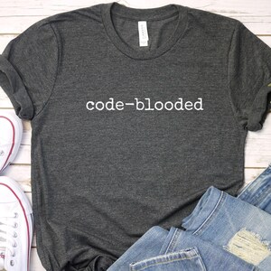 Peut inclure: T-shirt gris foncé avec l'inscription blanche "code-blooded". L'image présente également un jean bleu et des baskets en toile blanches. L'ensemble est posé sur une surface claire imitation bois.