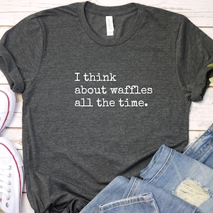 May include: Dark gray t-shirt with the text "I think about waffles all the time" in white. The shirt is paired with blue jeans and white canvas sneakers.