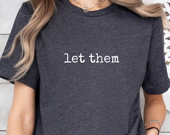 May include: Dark gray t-shirt with the text "let them" in white. The shirt has short sleeves and a crew neck. The fabric appears to be soft and comfortable. A simple, casual top.