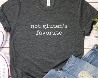 Puede incluir: Camiseta gris oscuro con el texto blanco "not gluten's favorite". Se combina con vaqueros azules y zapatillas blancas con detalles rojos. El conjunto informal está sobre una superficie de madera clara.