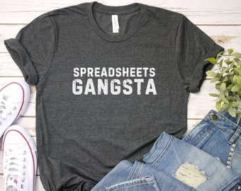 May include: A dark gray t-shirt with the text "SPREADSHEETS GANGSTA" printed in white on the front. The shirt is laid flat on a white wooden surface with a pair of white sneakers and a pair of blue jeans.