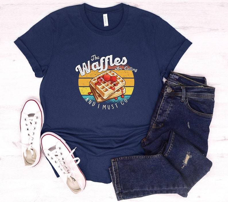 Op de afbeelding: Een marineblauw t-shirt met een vintage-stijl afbeelding van een wafel met aardbeien en de tekst "The Waffles Are Calling And I Must Go".