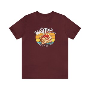 Op de afbeelding: Een maroon t-shirt met een retro-stijl grafische afbeelding van wafels met een rode bessen topping. De tekst luidt "The Waffles Are Calling And I Must Go."