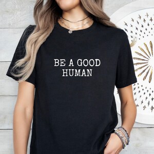 May include: Black t-shirt with the white text "BE A GOOD HUMAN". The shirt is a crew neck style and appears to be made of a soft material. The image shows the shirt on a person.