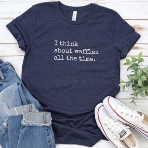 May include: Navy blue t-shirt with the text "I think about waffles all the time." The shirt is paired with blue jeans and white sneakers. A sprig of green leaves is visible in the background.