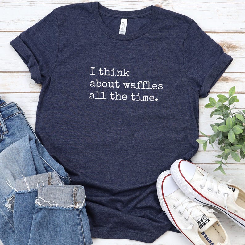May include: Navy blue t-shirt with the text "I think about waffles all the time." The shirt is paired with blue jeans and white sneakers. A sprig of green leaves is visible in the background.