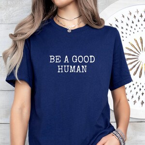 May include: Navy blue t-shirt with the white text "BE A GOOD HUMAN". The shirt is a short-sleeved crew neck style. The image shows the upper body of a person wearing the shirt.