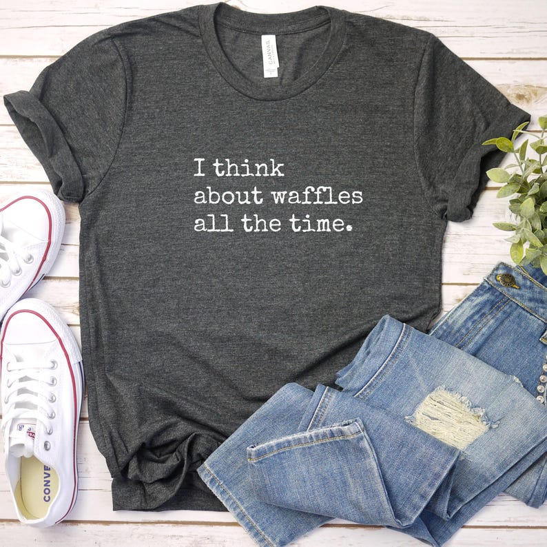 May include: Dark gray t-shirt with the text "I think about waffles all the time" in white. The shirt is paired with blue jeans and white canvas sneakers.