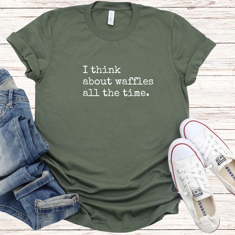 May include: Olive green t-shirt with rolled-up sleeves. The shirt features the text "I think about waffles all the time" in white. White canvas sneakers and blue jeans are visible in the image.