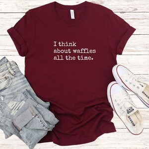 May include: A maroon t-shirt with the text "I think about waffles all the time." The shirt is paired with blue jeans and white sneakers. The shirt is made of a soft material and has rolled-up sleeves.