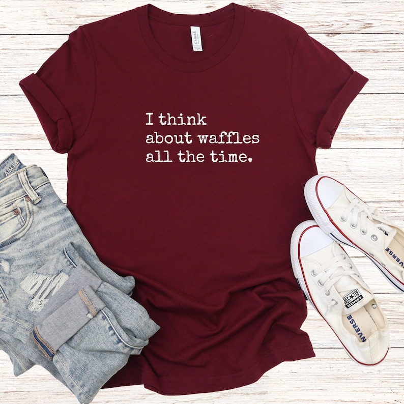 May include: A maroon t-shirt with the text "I think about waffles all the time." The shirt is paired with blue jeans and white sneakers. The shirt is made of a soft material and has rolled-up sleeves.