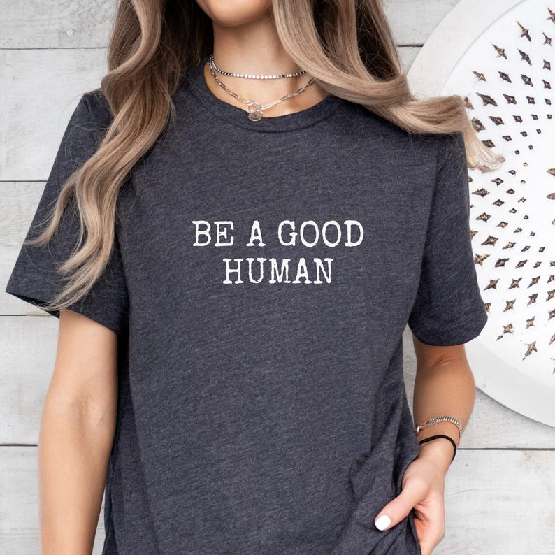 May include: Dark gray t-shirt with the white text "BE A GOOD HUMAN". The short-sleeved shirt is made of a soft material. The shirt is a casual wear item.