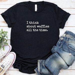 May include: Black t-shirt with the text "I think about waffles all the time" in white. The shirt is paired with blue jeans and white sneakers with red accents. The shirt is made of a soft material.