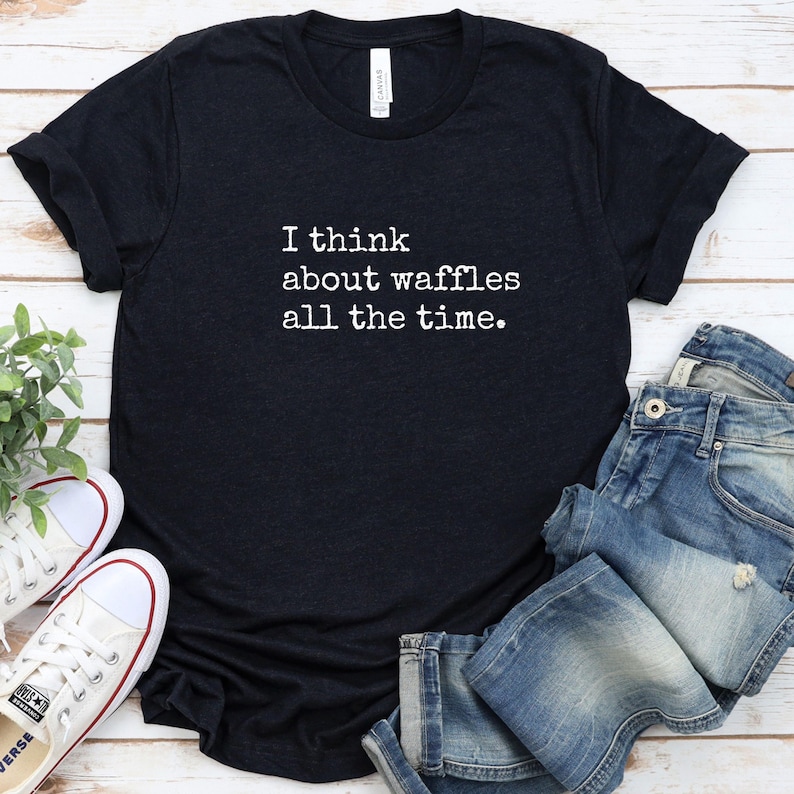 May include: Black t-shirt with the text "I think about waffles all the time" in white. The shirt is paired with blue jeans and white sneakers with red accents. The shirt is made of a soft material.