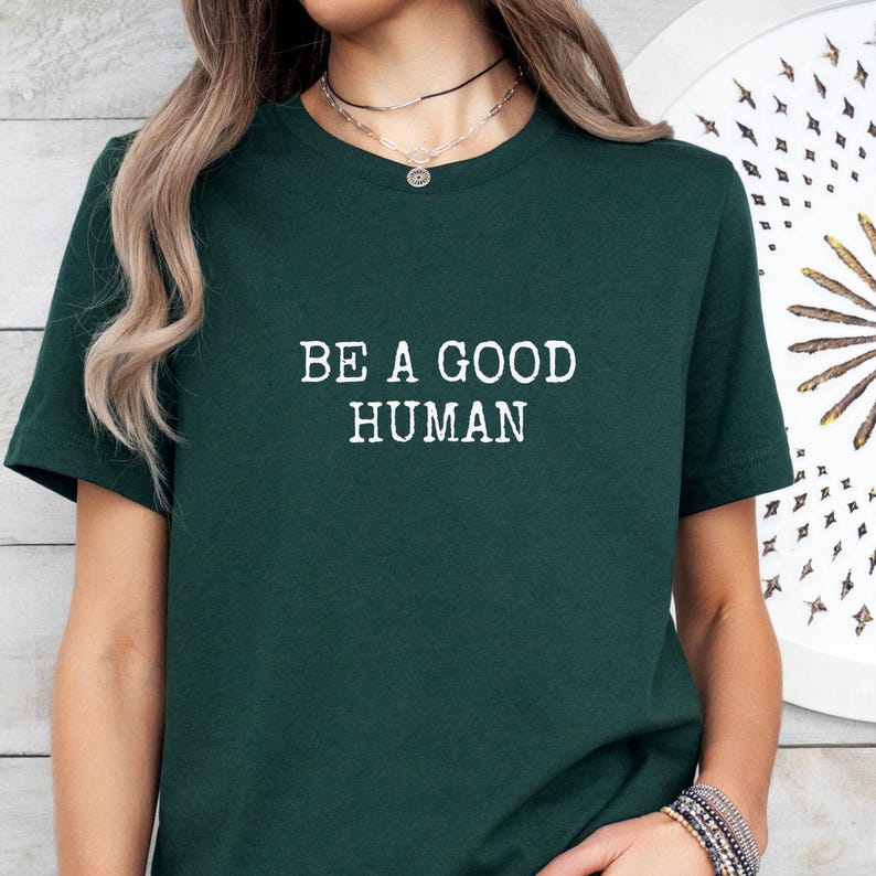 May include: A dark green t-shirt with the white text "BE A GOOD HUMAN" in a bold, sans-serif font. The shirt has short sleeves and a crew neck. The overall style is casual and comfortable.