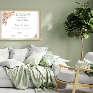 K&ouml;nnte beinhalten: Eine gerahmte Verpflichtungsurkunde mit den Namen Daniel Mars und Greta Mae Evans. Die Urkunde hat florale Akzente und wird &uuml;ber einem wei&szlig;en Sofa mit gr&uuml;nen Wurfkissen und einer Decke angezeigt. Rechts ist eine gro&szlig;e Topfpflanze.