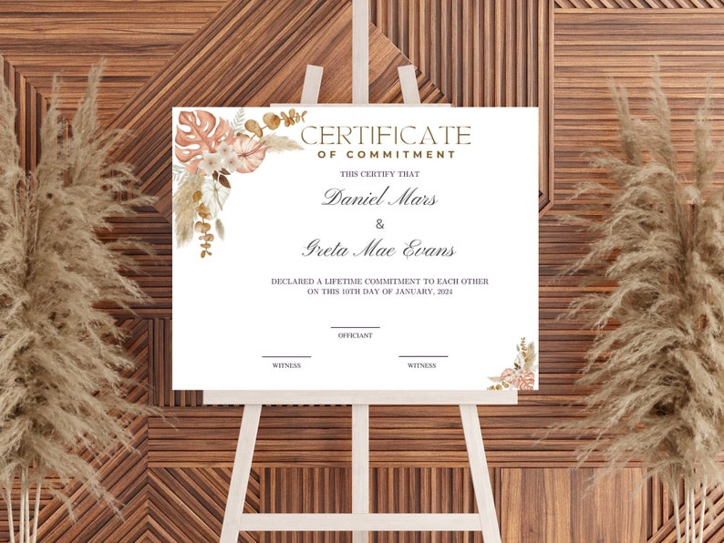 K&ouml;nnte beinhalten: Eine wei&szlig;e Verpflichtungsurkunde auf einer h&ouml;lzernen Staffelei. Die Urkunde tr&auml;gt die Aufschrift "CERTIFICATE OF COMMITMENT" und die Namen "Daniel Mars & Greta Mae Evans". Florale Akzente in Pfirsich und Braun schm&uuml;cken die Ecken. Der Hintergrund ist eine Holzwand.