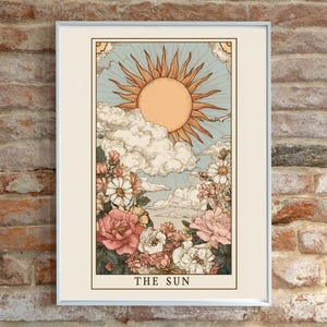 May include: Framed art print featuring a vintage-style illustration of the sun tarot card. The artwork depicts a radiant sun, clouds, and flowers in shades of orange, blue, and pink. The text "THE SUN" is at the bottom.