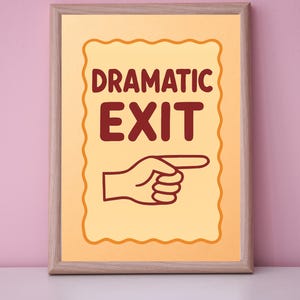 May include: A framed print with the words "DRAMATIC EXIT" in maroon letters. Below the text is a hand pointing to the right. The background is a light orange color with a decorative border. The frame is a light brown color.