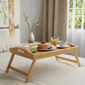 May include: A light brown bamboo breakfast tray with foldable legs, featuring a stack of pancakes topped with raspberries and blueberries, a small bowl of yogurt, a glass of orange juice, and a white cup of tea on a matching saucer.