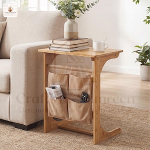 May include: A bamboo side table with a natural finish. It features a tabletop, a magazine rack with multiple pockets, and a C-shaped leg design. A newspaper and remote control are in the pockets. A stack of books and a mug sit on the table.